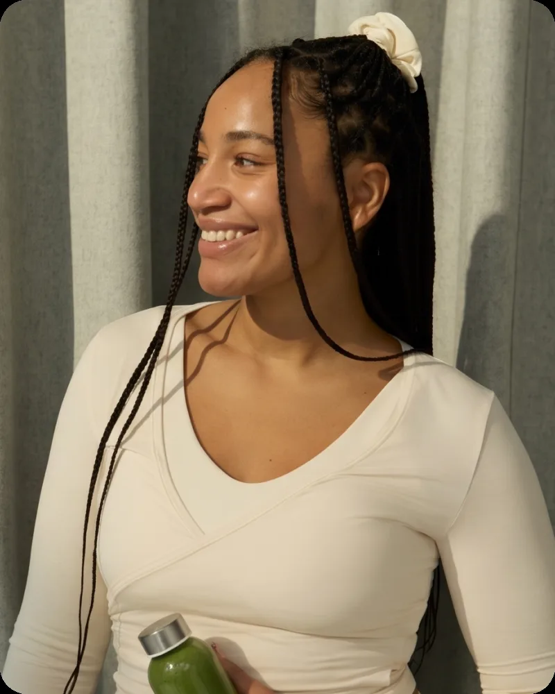 A smiling woman with long braids in a cream top, holding a green smoothie