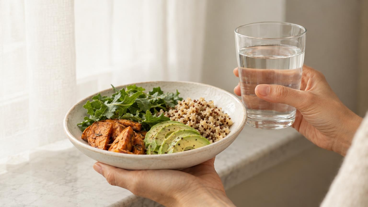 A plate of sliced avocado, leafy greens and quinoa beside a glass of water on a pale wooden table in soft daylight.